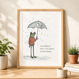 K&ouml;nnte beinhalten: Ein gerahmter Kunstdruck mit einem gr&uuml;nen Frosch in einem roten Pullover, der im Regen unter einem kaputten schwarzen Regenschirm steht. Der Text lautet, everything is okay if we ignore several things. Die Illustration ist in einem einfachen Stil.