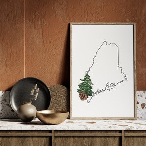 May include: A framed print of the outline of the state of Maine with a pine tree and pine cone in the lower left corner. The print is sitting on a countertop with a black plate, a woven placemat, a gold bowl, and a vase with dried grasses.