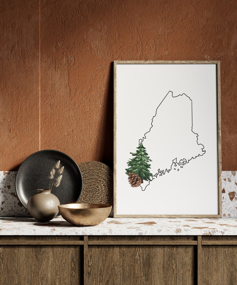 May include: A framed print of the outline of the state of Maine with a pine tree and pine cone in the lower left corner. The print is sitting on a countertop with a black plate, a woven placemat, a gold bowl, and a vase with dried grasses.
