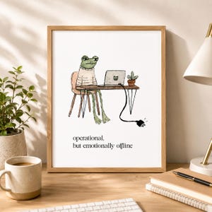 Könnte beinhalten: Ein gerahmter Kunstdruck mit einer Aquarellillustration eines grünen Frosches in einem beigen Pullover, der an einem Schreibtisch mit einem Laptop und einer kleinen Topfpflanze sitzt. Der Text lautet: "operational, but emotionally offline."
