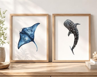 May include: Two framed wall art prints featuring watercolor illustrations of marine life. The left print displays a blue manta ray, while the right print shows a dark gray whale shark with white spots, both set against a plain white background.