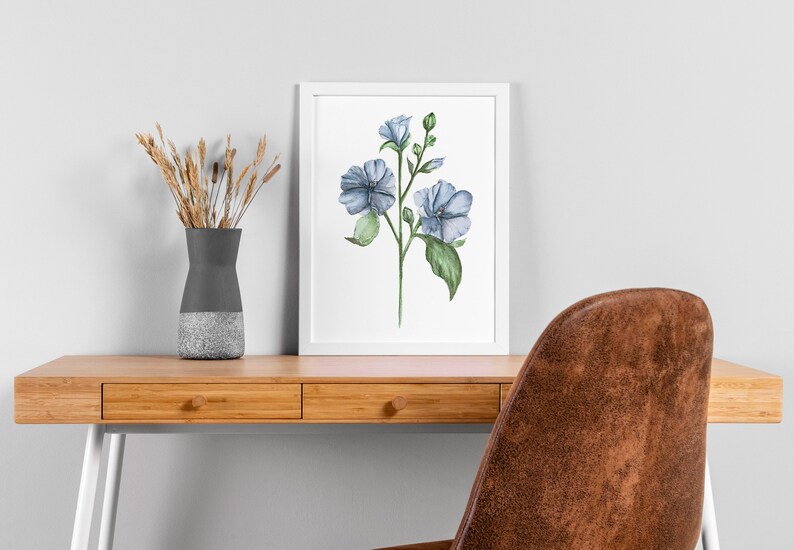 May include: A watercolor painting of blue flowers with green leaves and stems on a white background. The painting is framed in a white frame and is hanging on a white wall.