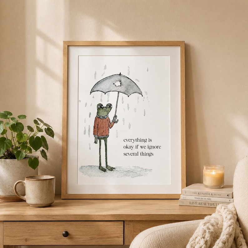 K&ouml;nnte beinhalten: Ein gerahmter Kunstdruck mit einer Aquarellillustration eines gr&uuml;nen Frosches in einem roten Pullover, der im Regen unter einem kaputten schwarzen Regenschirm steht. Der Text lautet, everything is okay if we ignore several things.