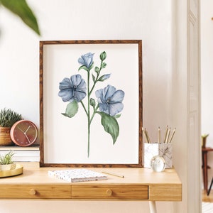 May include: A framed watercolor painting of a blue flower with green leaves on a white background. The painting is hanging on a wall above a wooden desk with a clock, pencils, and a notebook.
