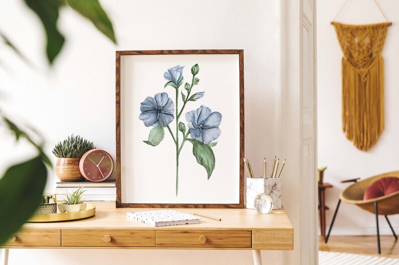 May include: A framed watercolor painting of a blue flower with green leaves on a white background. The painting is hanging on a wall above a wooden desk with a clock, pencils, and a notebook.