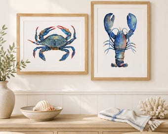 Może przedstawiać: Dwa oprawione wydruki ścienne przedstawiające akwarelowe ilustracje niebieskiego kraba i niebieskiego homara. Krab jest przedstawiony w orientacji poziomej, a homar pionowo, oba na gładkim, białym tle.