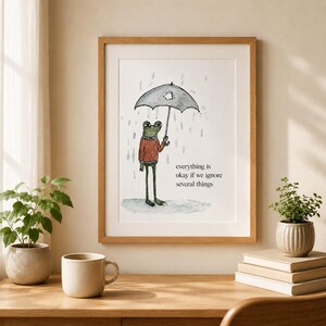 K&ouml;nnte beinhalten: Ein gerahmter Kunstdruck mit einer Aquarellillustration eines gr&uuml;nen Frosches in einem roten Pullover, der einen grauen Regenschirm im Regen h&auml;lt. Der Text lautet, everything is okay if we ignore several things. Der Druck befindet sich in einem hellen Holzrahmen.