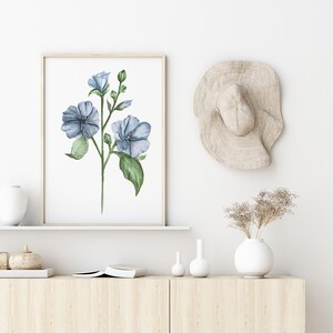 May include: A watercolor painting of blue flowers with green leaves in a light wood frame hanging on a white wall above a wooden shelf with a white vase and dried flowers, two small white vases, and a book.
