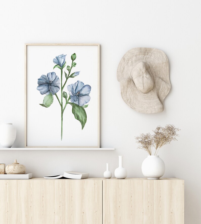 May include: A watercolor painting of blue flowers with green leaves in a light wood frame hanging on a white wall above a wooden shelf with a white vase and dried flowers, two small white vases, and a book.