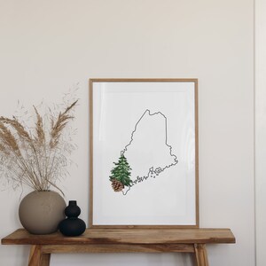 May include: A framed print of the outline of the state of Maine with a small pine tree and pine cone in the lower left corner. The print is hanging on a light wood shelf with a vase of dried pampas grass on the left.