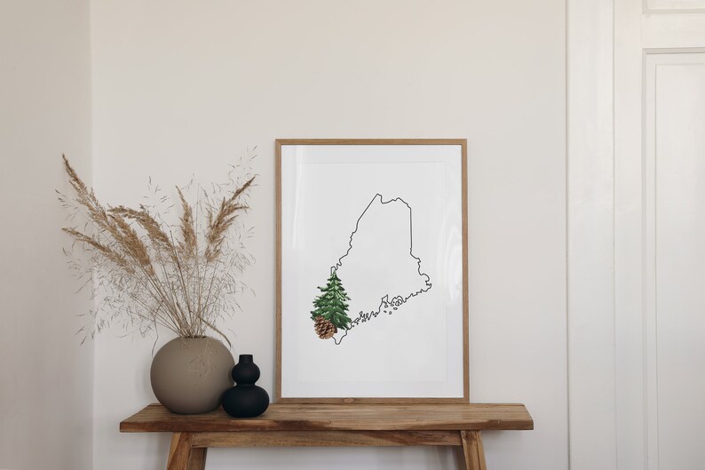 May include: A framed print of the outline of the state of Maine with a small pine tree and pine cone in the lower left corner. The print is hanging on a light wood shelf with a vase of dried pampas grass on the left.