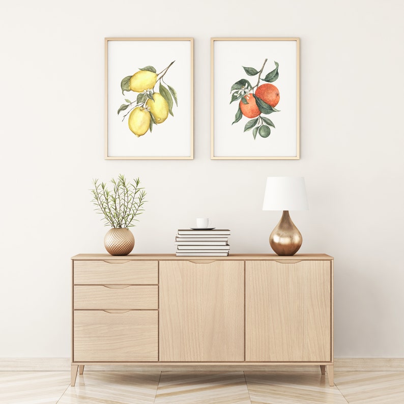 May include: Two framed watercolor prints of fruit branches. The left print features a lemon branch with yellow lemons and green leaves. The right print features an orange branch with orange fruit and green leaves. The prints are hanging on a white wall above a light wood dresser with two drawers and two doors. A gold vase with a small green plant sits on the dresser. A gold lamp with a white shade sits on the dresser.
