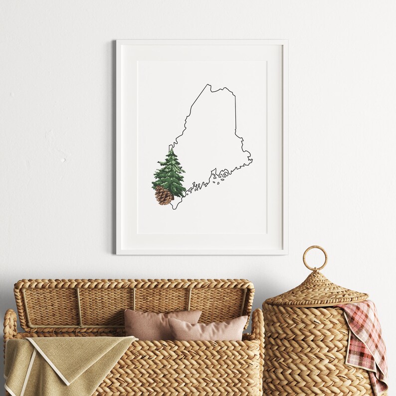 May include: A black and white line drawing of the state of Maine with a green pine tree and brown pine cone in the lower left corner. The print is framed and hanging on a white wall above a wicker basket.