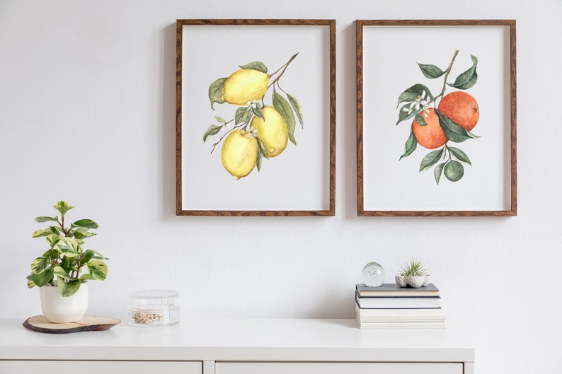 May include: Two framed watercolor paintings of fruit branches. The left painting shows a branch of lemons with green leaves. The right painting shows a branch of oranges with green leaves.