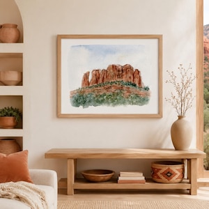 Könnte beinhalten: Ein gerahmtes Aquarell-Landschaftsbild, das rote Felsformationen vor einem blassen Himmel zeigt, mit grüner Vegetation an der Basis. Das Kunstwerk ist in einem hellen Holzrahmen gefasst und fängt die natürlichen Texturen und Farben der Szene ein.