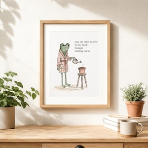 Könnte beinhalten: Ein gerahmter Kunstdruck mit einem grünen Frosch in einem rosafarbenen karierten Bademantel, der eine kleine Topfpflanze auf einem Holzhocker gießt. Der Text lautet: may the odds be ever in my favor because nothing else is.