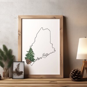 May include: A framed print of the state of Maine with a pine tree and pine cone in the lower left corner. The print is on a white background and is framed in a natural wood frame. The print is sitting on a wooden table with a pine branch, a pine cone, and a lamp on the table.