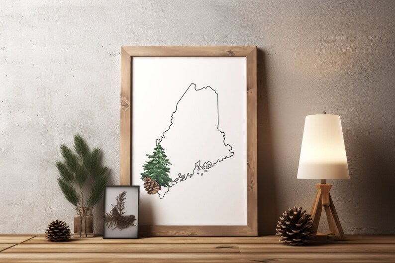 May include: A framed print of the state of Maine with a pine tree and pine cone in the lower left corner. The print is on a white background and is framed in a natural wood frame. The print is sitting on a wooden table with a pine branch, a pine cone, and a lamp on the table.