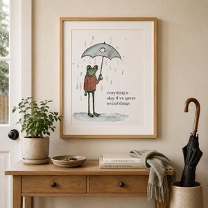K&ouml;nnte beinhalten: Ein gerahmter Kunstdruck mit einer Aquarellillustration eines gr&uuml;nen Frosches in einem roten Pullover, der einen zerrissenen Regenschirm im Regen h&auml;lt. Der Text lautet, everything is okay if we ignore several things, in schwarzer Schrift.