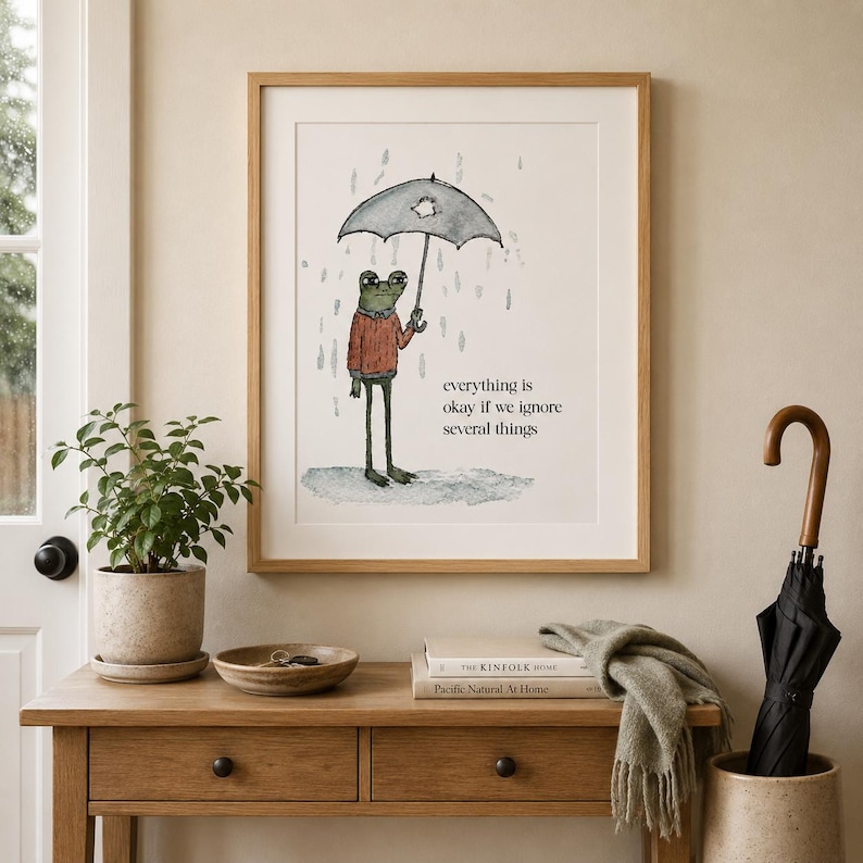 K&ouml;nnte beinhalten: Ein gerahmter Kunstdruck mit einer Aquarellillustration eines gr&uuml;nen Frosches in einem roten Pullover, der einen zerrissenen Regenschirm im Regen h&auml;lt. Der Text lautet, everything is okay if we ignore several things, in schwarzer Schrift.