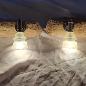 May include: Two rustic light fixtures mounted on weathered wood. Each fixture features a clear glass insulator shade and a black metal pipe fitting. The lights are illuminated, casting a warm glow. The wood has a natural, aged appearance.
