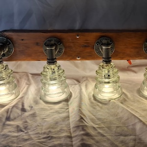 May include: A rustic, handmade light fixture featuring four clear glass insulator shades. The shades are attached to a wooden beam with metal pipe fittings. The lights are illuminated, casting a warm glow.