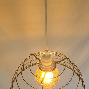 May include: A gold-toned metal cage pendant light fixture with a clear light bulb. The light is hanging from a white cord.