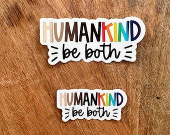 May include: Two white stickers with the text "HUMANKIND be both". The word "HUMANKIND" is in a variety of skin tone colors and rainbow colors. The stickers are on a wooden surface.