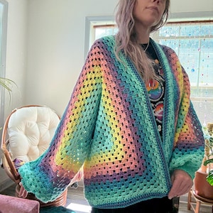May include: A handmade, open-front crochet cardigan in a rainbow of colors, including yellow, pink, blue, and green. The cardigan features a loose, open weave and long sleeves. The garment is a vibrant and colorful piece of clothing.