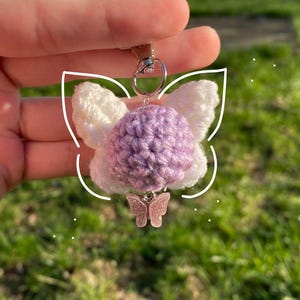 May include: A handmade keychain featuring a crochet fairy with white wings and a lavender body. A small pink butterfly charm hangs below. The keychain is held against a blurred green background.