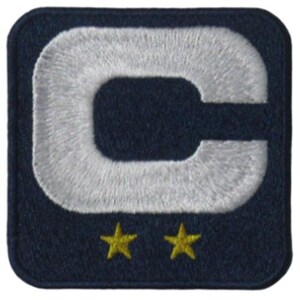 May include: Embroidered patch with a navy blue background and a large, white, stylized letter "C". Two small, yellow stars are positioned below the letter. The patch has a square shape with rounded corners.