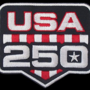 May include: A rectangular embroidered patch with rounded corners and a pointed bottom edge. The patch features the white text USA above the number 250, which includes a small white star. Red and white vertical stripes accent the design.