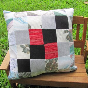 May include: A patchwork pillow with a checkered pattern in red, black, white, and gray. The pillow is made of different fabrics and has a variety of textures. It is sitting on a wooden bench.