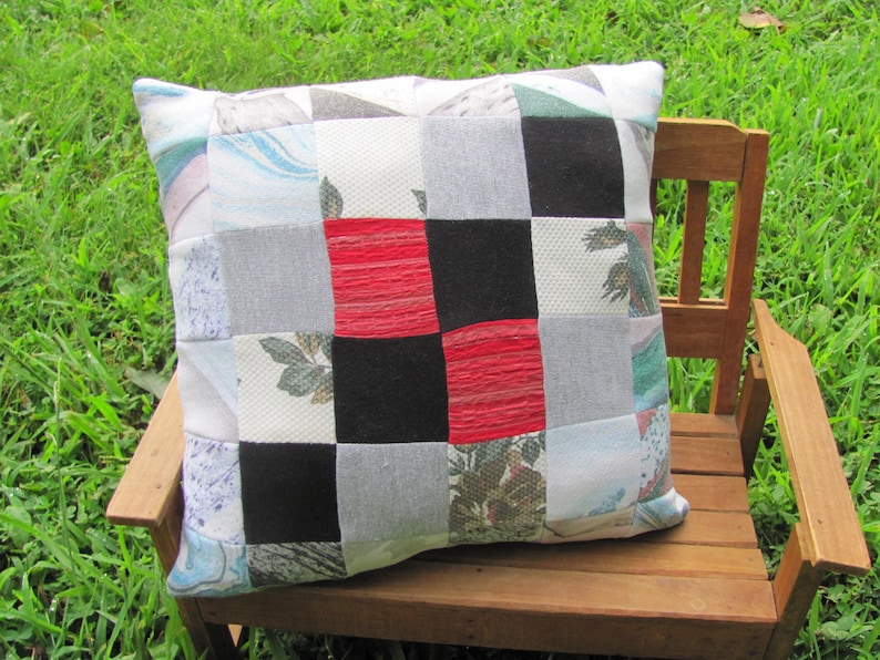 May include: A patchwork pillow with a checkered pattern in red, black, white, and gray. The pillow is made of different fabrics and has a variety of textures. It is sitting on a wooden bench.
