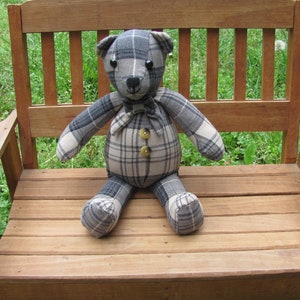 May include: A plaid teddy bear with black button eyes sits on a wooden bench. The bear is made from a patchwork of plaid fabric in shades of gray and white. The bear has a bow tie and two large buttons on its chest.