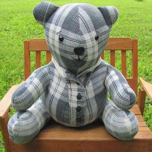 May include: A handmade teddy bear made of blue, white, and red plaid fabric. The bear is wearing a collared shirt with three dark blue buttons and a white collar. It is sitting on a wooden chair with a green grassy background.