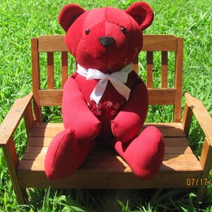 May include: A handmade red teddy bear with a white bow tie sits on a small wooden bench. The bear has a corduroy texture on its head and a floral pattern on its chest. The bench is made of wood and is surrounded by green grass.