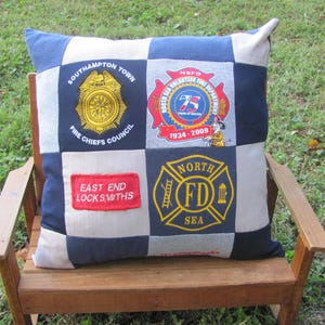 May include: A patchwork pillow with four different patches featuring logos for a fire department, a fire chiefs council, a volunteer fire department, and a locksmith company. The pillow is on a wooden bench.