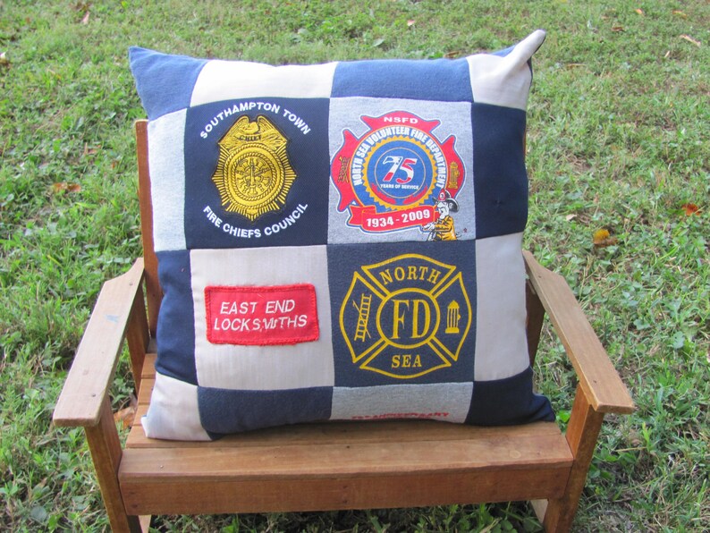 May include: A patchwork pillow with four different patches featuring logos for a fire department, a fire chiefs council, a volunteer fire department, and a locksmith company. The pillow is on a wooden bench.