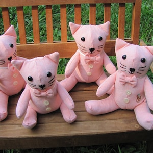 May include: Four pink plush cat toys with black eyes, black whiskers, and black embroidered mouths. Each cat is wearing a white bow tie and has a button on its chest. The cats are sitting on a wooden bench.