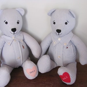May include: Two blue and white striped teddy bears with embroidered hearts on their feet. The bears are wearing shirts with collars and buttons. The heart on the left bear says "GHL3" and the heart on the right bear says "GHLO".