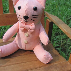 May include: A pink plush cat toy with a bow tie and button details. The cat is sitting on a wooden chair.