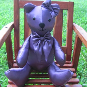 May include: A stuffed teddy bear made of smooth, shiny purple fabric. The bear features a large purple bow tie at the neck, a matching bow on the top of the head, round black button eyes, and a stitched black nose and mouth on its face.
