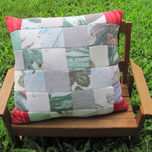 May include: A patchwork pillow with a checkered pattern of various shades of gray, white, and green. The pillow has red trim and is sitting on a wooden bench.