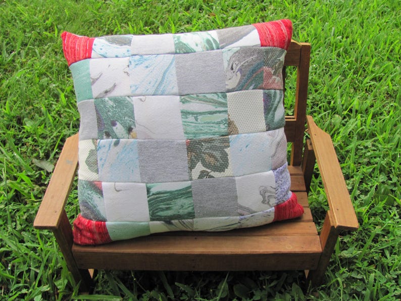 May include: A patchwork pillow with a checkered pattern of various shades of gray, white, and green. The pillow has red trim and is sitting on a wooden bench.