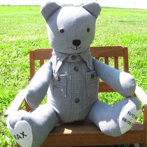 May include: A gray denim teddy bear wearing a denim shirt with buttons and pockets. The bear is sitting on a wooden bench with the text "MAX" on one foot and "11/17/11 08/06/12" on the other foot.