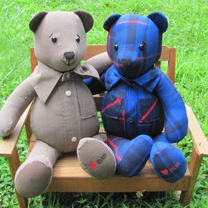 May include: Two plush teddy bears wearing shirts, sitting on a wooden bench. The bear on the left is wearing a gray and white striped shirt, and the bear on the right is wearing a blue and red plaid shirt. The bear on the left has "DAD" embroidered on its foot, and the bear on the right has "MOM" embroidered on its foot.
