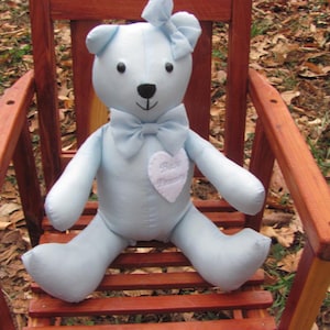 May include: A light blue teddy bear with a bow on its head and a bow tie. The bear has a white heart-shaped patch on its chest with the words "Baby Dreams" embroidered on it. The bear is sitting on a wooden chair.