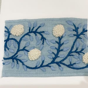 May include: A blue and white embroidered fabric panel with a floral design. The flowers are white and puffy, and the leaves are blue. The fabric is a light blue linen.