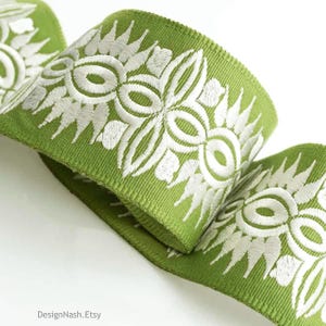May include: Close-up of a green ribbon with white floral embroidery. The ribbon is folded over itself, showcasing the detailed stitching and texture. The design features a repeating pattern of stylized flowers and leaves.